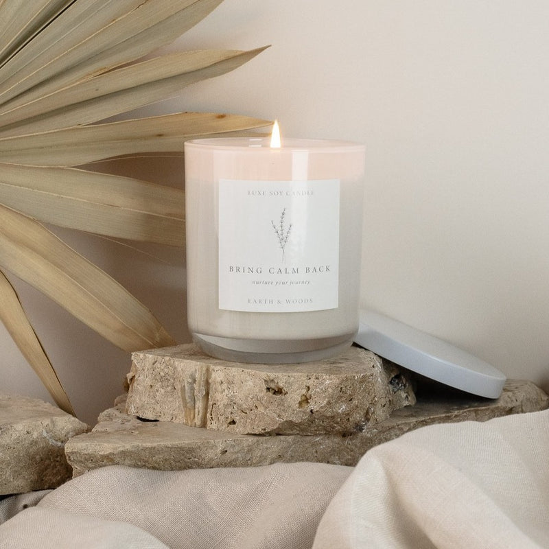 Earth and Woods candle in a glass jar with a sealable lid and drawstring bag. The candle's warm and familiar scent features notes of patchouli, sandalwood, vanilla, bergamot, lemon peel, cedarwood, and white musk. Burn time of 40+ hours.