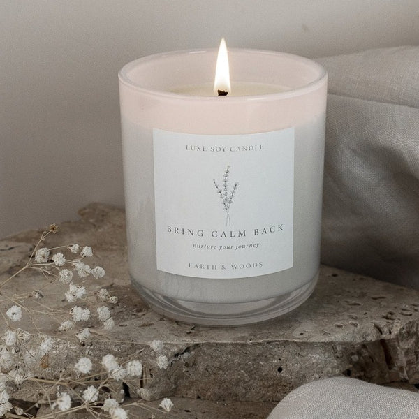 Earth and Woods candle in a glass jar with a sealable lid and drawstring bag. The candle's warm and familiar scent features notes of patchouli, sandalwood, vanilla, bergamot, lemon peel, cedarwood, and white musk. Burn time of 40+ hours.